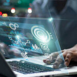 Person's arm/hand typing on laptop with computer graphics over keyboard of gears and symbols