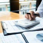 Hand of person holding a pen and typing on a calculator on top of a clipboard
