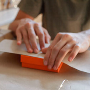 Person wrapping small orange box with wrapping paper and additional organge gift boxes stacked up