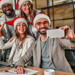 Happy New Year and Merry Christmas! Multiracial young creative people in Santa hats celebrate a holiday in a modern office and take a selfie with a smartphone on their last working day.