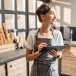 Woman in an apron working on a laptop standing in front of a cabinet with rulers on top of it