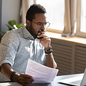 Focused serious african american businessman accountant analyst holding documents looking at laptop computer screen doing online trade market tech research thinking working sit at home office desk