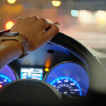 Close up of driver hands holding steering wheel driving car with blurred city street lights on background at night