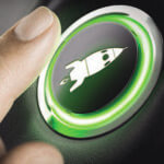 A person's finger about to push a lit button with an image of a rocket ship