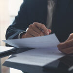 Signing contract, business agreement and deal concept. Closeup businessman in black suit reading before signing business contract documents at desk with laptop computer, busy with paperwork. Man manager proofing report at office
