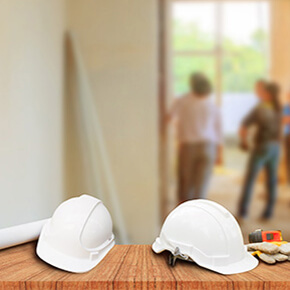 Two white hardhats sitting on a work bench with 2 people standing in the background talking