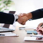 Two business men shaking hands at a desk full of paperwork