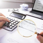 Hand using calculator, magnifying glass, glasses laptop on table looking closely at paper
