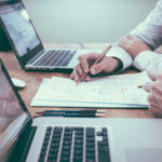 Two partial views of people working on papers and two laptops on a desk near them