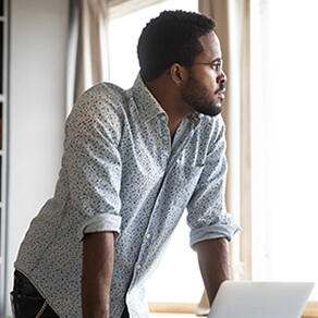 Man resting his arms on a desk in front of a laptop looking out a window