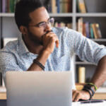 Man sitting in front of a laptop resting his fist on his chin and looking to his left concerned