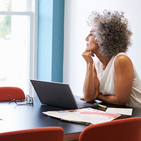 Lady sitting at a table with laptop, papers and glasses looking out a window in deep thoughts
