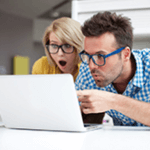 Lady with glasses and man with blue glasses looking happily surprised at something on a laptop